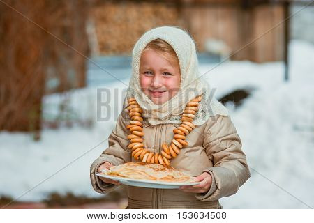 Sweet girl in old Russian scarf on the head with lengths in a plate and bread-rings around the neck walking down the street