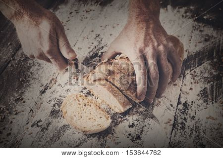 Baking and cooking concept background. Hands of baker cutting bread loaf with knife on rustic wooden table sprinkled with flour. Stained dirty hands of baker
