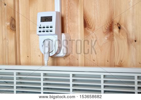 Working white electric convector heater plugged to timer power socket operated in smart house system against wooden wall inside room side view closeup