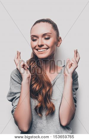 Waiting for special moment. Attractive young smiling woman keeping her fingers crossed and eyes closed while standing against grey background