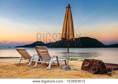 Summer Vacation end concept. Sunset tropical beach with two old chairs and a folded umbrella. Landscape with a quiet sea and the mountain in the distance Koh Chang Thailand.