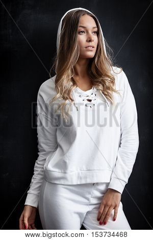 Young trendy woman in white sportsuit posing near black wall