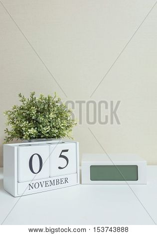 White wooden calendar with black 5 november word with clock and plant on white wood desk and cream wallpaper textured background selective focus at the calendar
