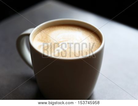 Cup of tasty latte is standing on the grey textured table. There is nice picture is painted on the top of the latte. Latte is in the white big cup.
