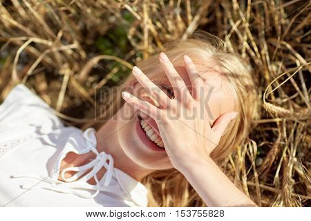 nature, summer holidays, vacation and people concept - close up of happy young woman lying on cereal field and covering face by hand