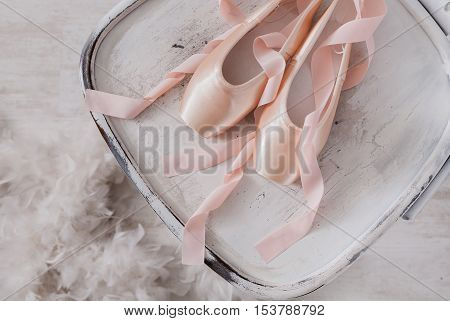 Pastel pink ballet shoes background. New pointe shoes with satin ribbon lay on white rustic shubby chic chair near feather boa, top view from above