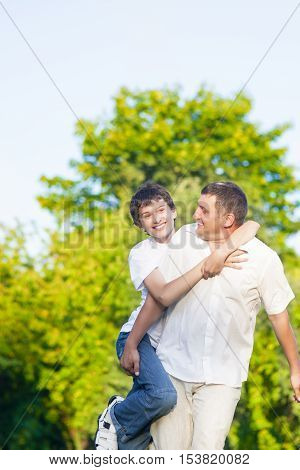 Happy Caucasian Family of Father and Son Piggybacking Outdoors. Against Nature Green Forest.Vertical Composition