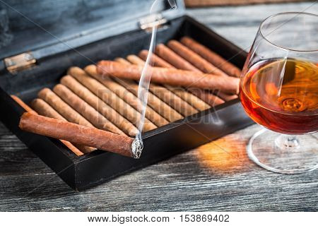 Burning cigar on humidor and cognac on old wooden table
