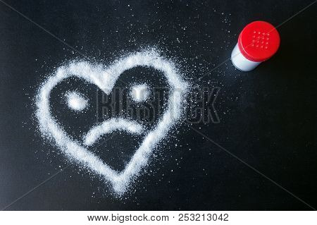 Salt Scattered On Black Surface. Drawn Heart With A Sad Face. Concept- Diet, Harm To Health From Exc