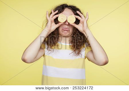 Curly Haired Funny Woman Closes Eyes With Two Halves Of Lemons And Opens Mouth Demonstrating Sour Ta