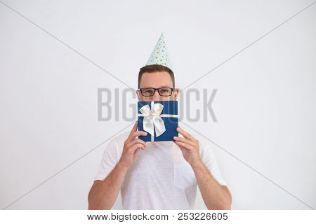 Young Man In Party Hat Covering Mouth With Gift Box. Handsome Man In Glasses Looking At Camera And H