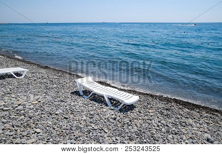 Chaise Lounge Is White On A Stony Beach By The Coastline. Chaise Lounge By The Sea.