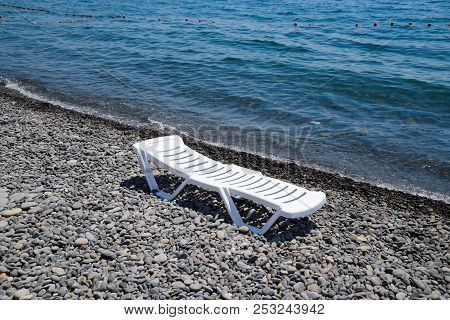 Chaise Lounge Is White On A Stony Beach By The Coastline. Chaise Lounge By The Sea.