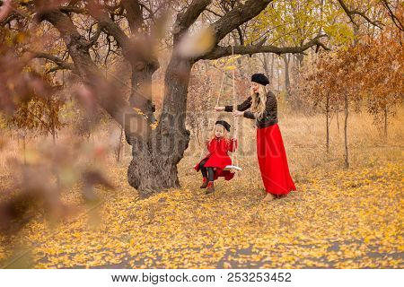 Beautiful Mother In A Long Dress Is Swinging On A Hinged Swing Little Daughter In A Red Coat In An A