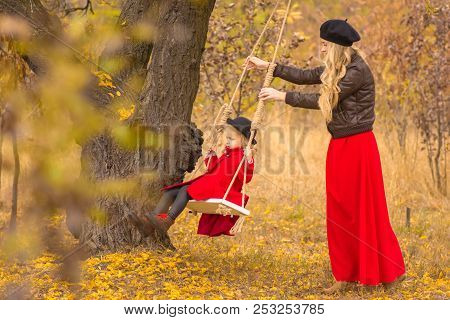 Beautiful Mother In A Long Dress Is Swinging On A Hinged Swing Little Daughter In A Red Coat In An A