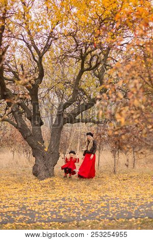 Beautiful Mother In A Long Dress Is Swinging On A Hinged Swing Little Daughter In A Red Coat In An A