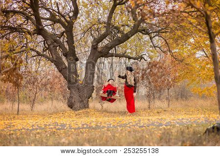 Beautiful Mother In A Long Dress Is Swinging On A Hinged Swing Little Daughter In A Red Coat In An A