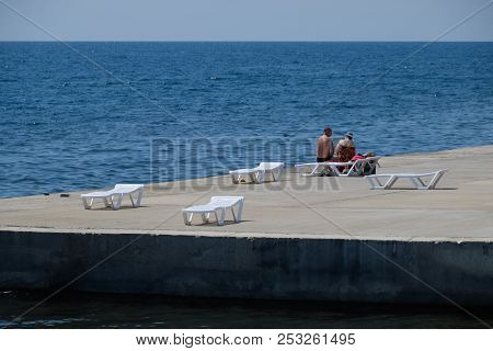 Novorossiysk, Russia - August 06, 2018: Chaise Lounge Is White On A Stony Beach By The Coastline. Ch