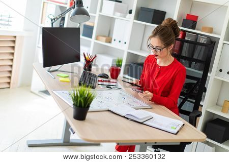 A Young Girl Is Sitting At The Desk In The Office, Holding A Bank Card And Phone In Her Hand.