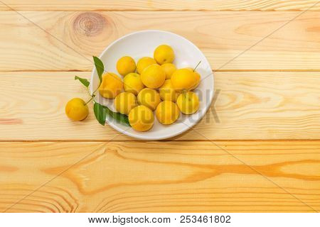 Yellow Cherry Plums On A White Saucer And One Fruit With Leaves Beside On A Light Colored Wooden Sur