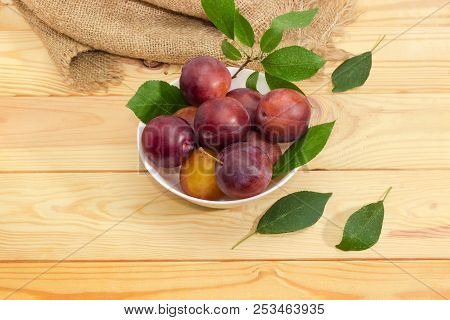 Ripe Purple Plums In White Bowl And Leaves Of Plum Tree On A Light Colored Wooden Surface