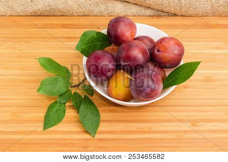 Ripe Purple Plums In White Bowl And Leaves Of Plum Tree On A Bamboo Wooden Surface