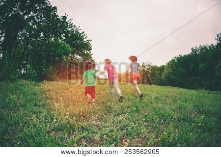 A Woman With Children Runs Along The Green Grass. Mom Holds Her Sons By The Hands And Run. A Boy Pla