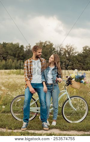 Smiling Lovers With Retro Bicycle Looking At Each Other In Field With Wild Flowers