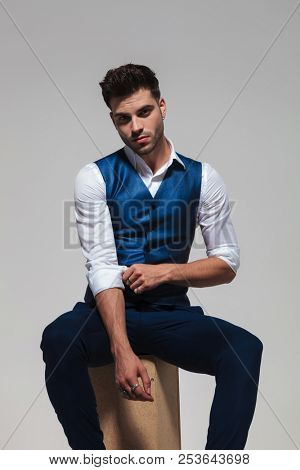 seated elegant man wearing a blue vest fixes his shirt sleeves on light grey background