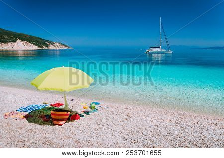 Umbrella And Beach Accessories On Pebble Beach On Lazy Summer Day. Sailing Boat Stays At Anchor In C