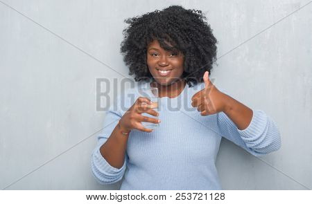 Young african american woman over grey grunge wall drinking a glass of water happy with big smile doing ok sign, thumb up with fingers, excellent sign