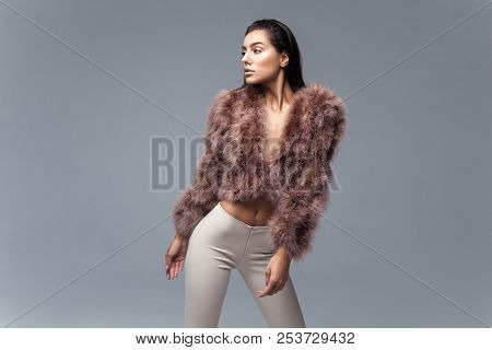 Young Elegant Girl Posing In Fluffy Pink Cape With Artificial Fur And Beige Flare Trousers On Gray B