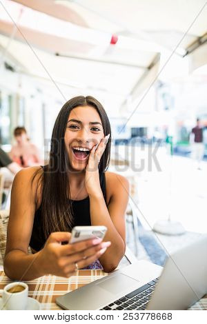 Young Latin Woman Getting Shocked By News In Her Phone Outdoors In Cafe. Shocked Face Of A Girl Sitt