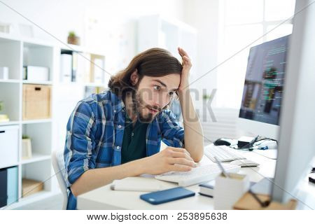 Young confused it-manager looking at one of computer monitors with tense expression