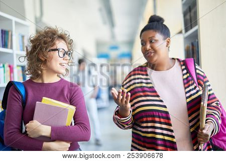 Positive friendly interracial students in bright clothing chatting while walking over corridor: confident African-American girl gesturing while explaining her idea to groupmate
