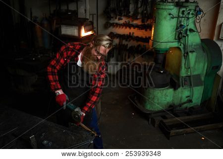 Serious concentrated brutal bearded worker in apron cutting metal with grinder while working in dark smithery