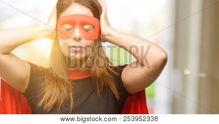 Middle age super hero woman wearing red mask and cape covering ears ignoring annoying loud noise, plugs ears to avoid hearing sound. Noisy music is a problem.