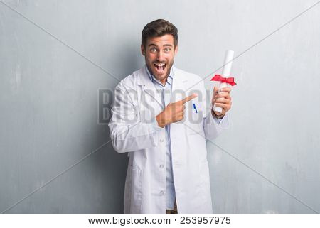 Handsome young doctor man over grey grunge wall holding diploma very happy pointing with hand and finger