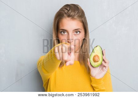 Beautiful young woman over grunge grey wall eating avocado pointing with finger to the camera and to you, hand sign, positive and confident gesture from the front
