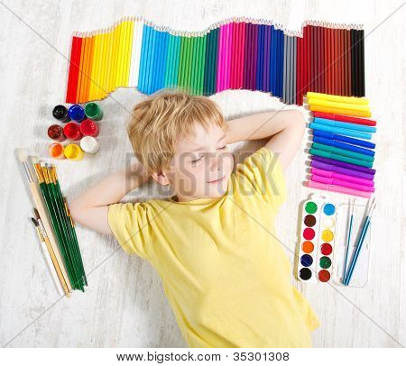 Bambino sognando sdraiato accanto a matite, pennelli e pitture.  Concetto di ispirazione e creatività