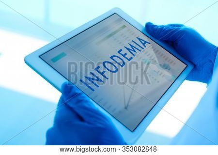 closeup of a doctor man, wearing blue surgical gloves, having a digital tablet in his hands with the word infodemia, infodemic written in spanish, in its screen
