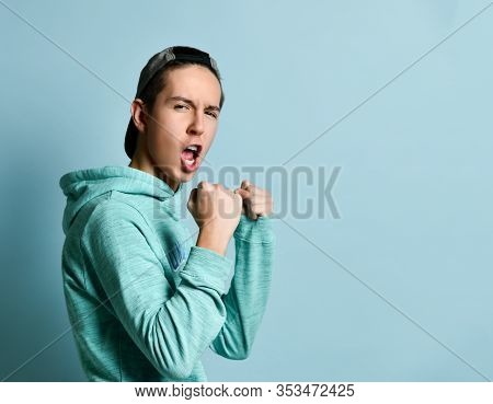 Young Teen Boy In Blue Hoodie Standing, Shouting And Ready To Defend Himself With Fists Over Pastel 