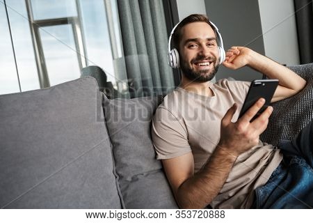 Image of a smiling optimistic young handsome man indoors at home on sofa listening music with headphones using mobile phone.