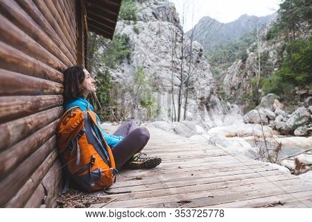 A Woman With A Backpack Sits On The Porch Of An Old Wooden House. The Traveler Found A Hut For An Ov