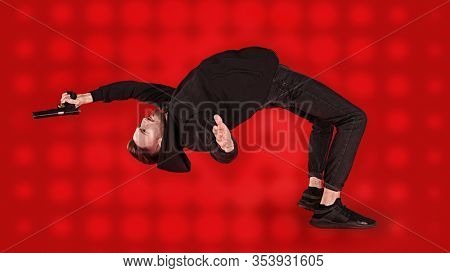 Handsome man in black clothes in motion in flight with a gun. Super agent on a mission. Criminal world, counterterrorism. Studio shot on a red background.
