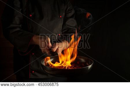 Professional Chef Prepares Vegetable Food In A Frying Pan With Flame Of Fire. The Concept Of Restaur