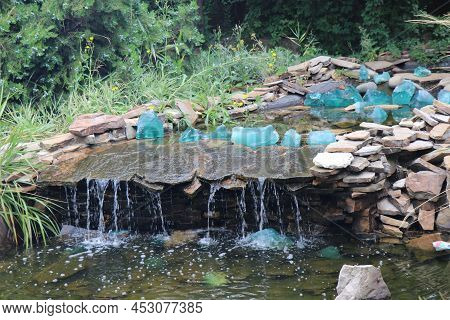 Beautiful Fountain With Rocks And Waterfalls. Fountain In The Park Near Big Green Trees