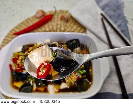 Detail Of Spoon With Piece Of Silken Tofu, Thousand Years Old Egg, And Chilli Pepper With Whole Dish