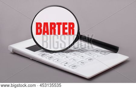 On A Gray Background, A White Calculator And A Magnifying Glass With The Text Barter. Business Conce
