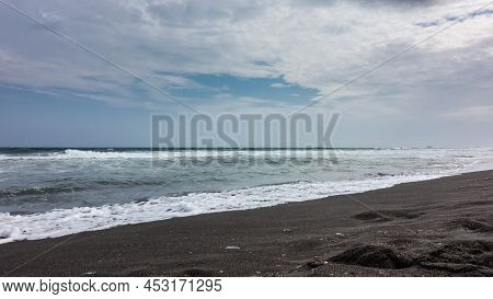 Long Waves Roll And Foam On The Beach With Black Volcanic Sand. Clouds In The Blue Sky. Kamchatka. P